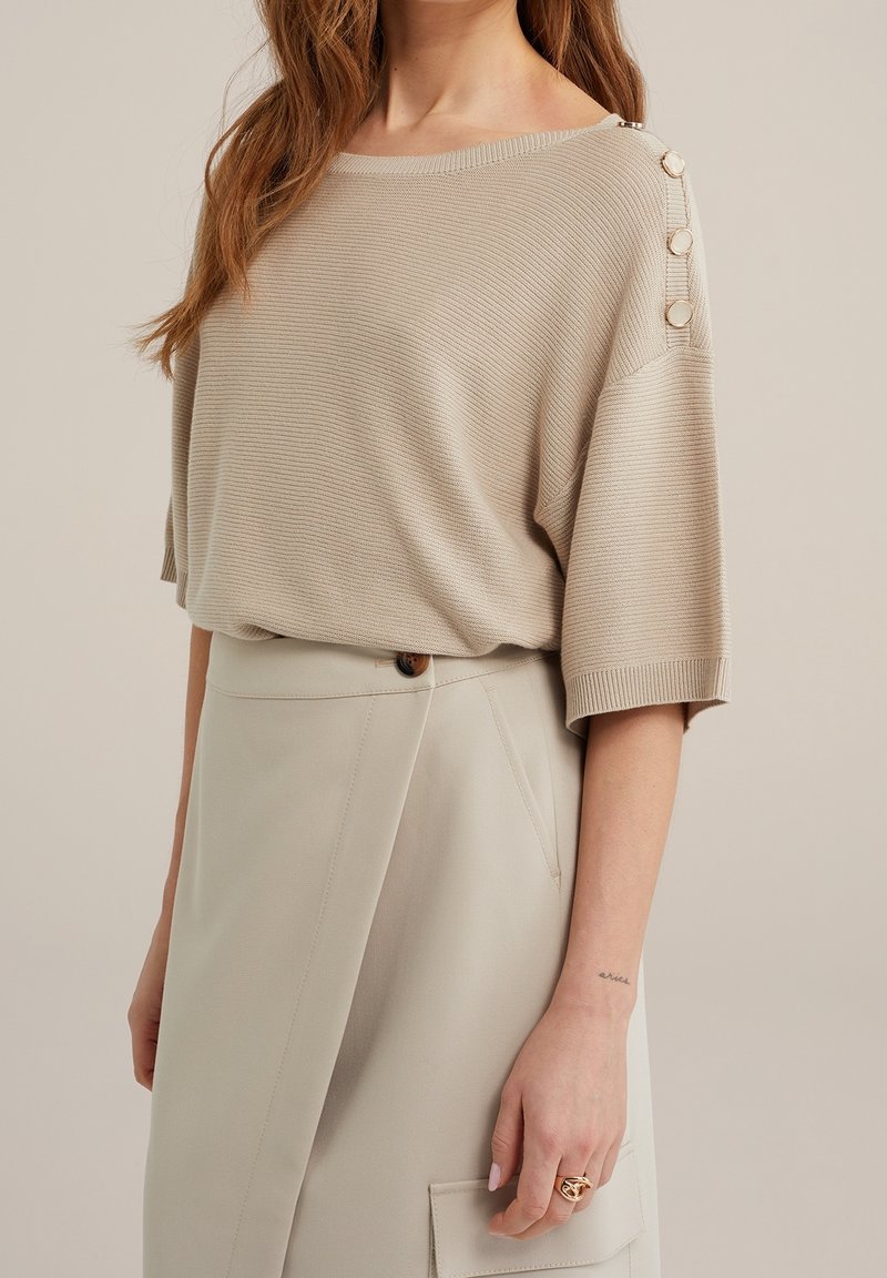 Beige stickad topp med korta ärmar och ribbad textur, med dekorrade knappar på axeln. Matchad med en ljusbeige kjol.