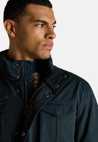 Muž v tmavomodrej bunde so zipsom a cvokmi sa pozerá na stranu, pričom ukazuje na detailný golier bundy a vrecko.