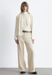 Beige long-sleeve top with a high collar paired with cream-colored corduroy wide-leg pants and a black belt. Smooth textures, minimal design.