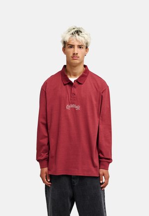 Jeune homme aux cheveux blonds portant un polo rouge à manches longues et un pantalon foncé, debout devant un fond clair uni.