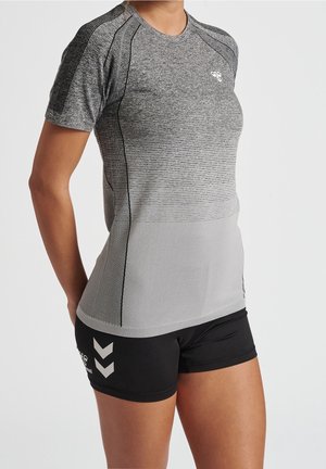Sportowiec ubrany w dopasowaną szarą sportową koszulkę z krótkim rękawem i czarne krótkie spodenki sportowe stoi na jednolitym tle.