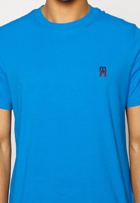 Blå bomulds-T-shirt med rund halsudskæring og korte ærmer. Markant logo i rød og blå på venstre bryst. Blød, glat tekstur.