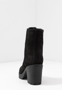 Stivaletto alla caviglia in suede nero con tacco a blocco spesso e zip sul retro, mostrato da un'angolazione posteriore su superficie e sfondo bianchi.