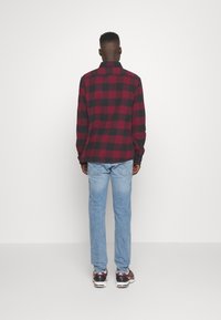 Camicia a quadri rossa e nera con maniche lunghe, polsini con bottoni e colletto. Indossata con jeans azzurri chiari e scarpe da ginnastica.