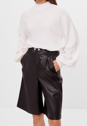 Kvinde iført hvid langærmet sweater og sorte læderknælange shorts med høj talje og lommer, stående med hænderne i lommerne.