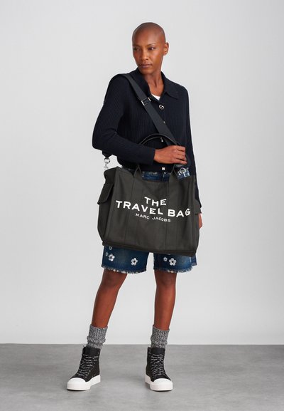 Saco de viagem preto em lona com texto branco que diz "THE TRAVEL BAG MARC JACOBS", apresentando alças robustas e uma alça de ombro ajustável.
