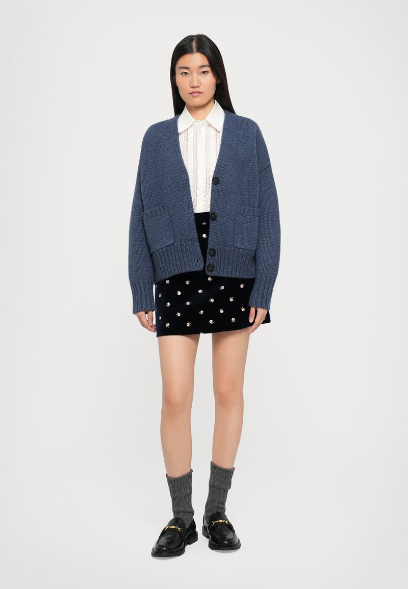 Jauna sieviete valkā zilu pārāk lielu kardigānu, baltu mežģīņu blūzi, melnu ziedu svārku, pelēkas zeķes un melnas loafer kurpes, stāvot pret neizteiksmīgu fonu.