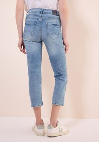 Jeans en denim bleu clair, coupés et à texture délavée, avec deux poches arrière, associés à des baskets blanches avec des accents verts.