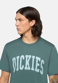 Tmavě zelené tričko vyrobené z jemné látky, na přední straně s grafickým textem "DICKIES" v světle modré barvě a tučném písmu. Krátké rukávy a kulatý výstřih.