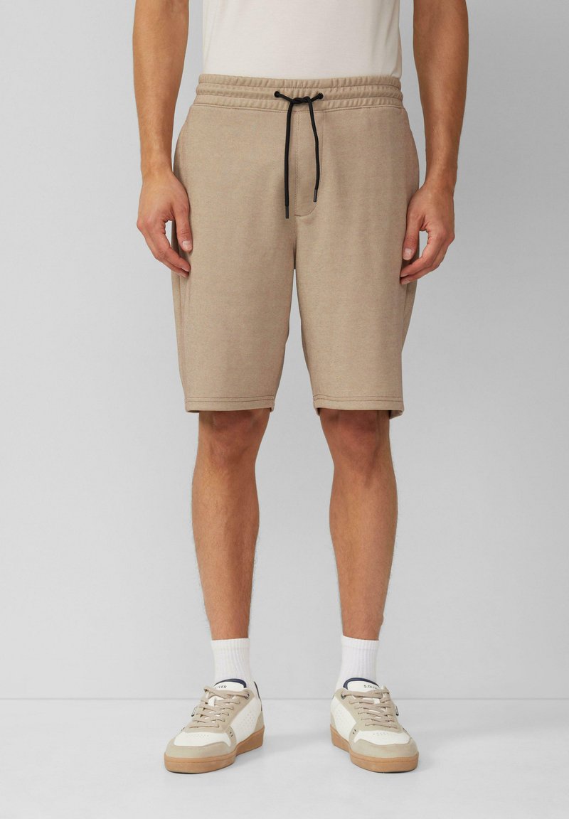 Beige Strickshorts mit elastischem Bund und schwarzem Kordelzug. Glatte Textur, knielang, kombiniert mit hellen Sneakers und weißen Socken.