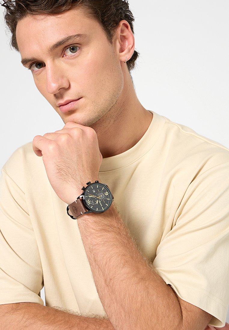 Reloj negro y dorado con una esfera redonda, números árabes, correa de cuero marrón, sobre un fondo de camiseta neutro.