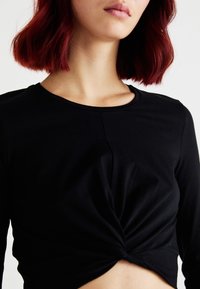 Zwarte langslieftop met een gedraaide voorkant. Heeft een ronde hals en zachte stof, dicht aansluitend bij het lichaam.