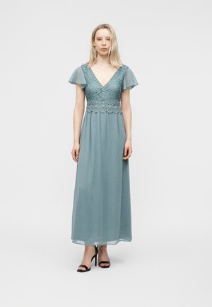 Vrouw staat in een golflange lichtblauwe jurk met een kanten lijfje, zwierige mouwen, een diepe V-hals en zwarte sandalen met hoge hakken.