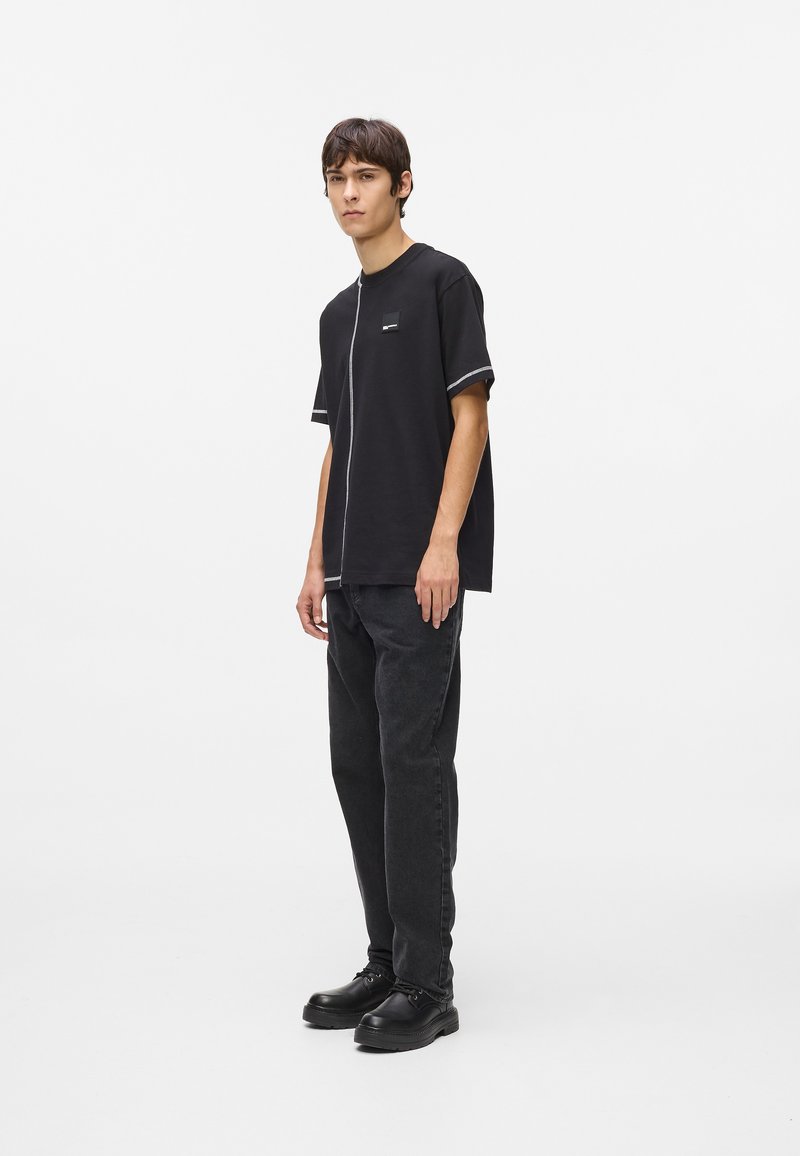 Camiseta negra de manga corta con costuras en contraste, que presenta un pequeño parche con logo, combinada con jeans gris oscuro y zapatos negros.