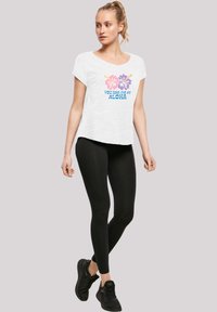 Weißes T-Shirt mit kurzen Ärmeln, das ein buntes Blumenmuster und den Text "DU HAST MICH BEI ALOHA GEHABT" zeigt. Kombiniert mit schwarzen Leggings und Sneakers.