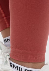 Leggings rouges en tissu lisse, dotés d'une coupe ajustée, de coutures sans couture et d'un petit logo au niveau de la cheville. Des chaussettes blanches visibles en dessous.