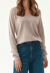 Suéter de punto de color beige claro, de manga larga y con escote en V, acompañado de unos vaqueros de tiro alto en azul oscuro. Textura suave, corte holgado y costuras detalladas.