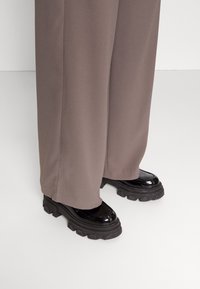 Svarta lacklädersplattformsskor med kraftiga gummisål, som bärs med löst sittande, ljusbruna byxor. Slät yta och glänsande finish.