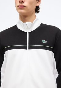 Άνδρας που φοράει ένα μαύρο και άσπρο μπουφάν με φερμουάρ και ψηλό γιακά, με πράσινο λογότυπο κροκοδείλου στο στήθος.