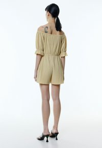 Schulterfreier, gelber, gefälteter Jumpsuit mit kurzen Ärmeln und tailliertem Gürtel. Kombiniert mit schwarzen, hohen Sandalen. Glatte Textur.