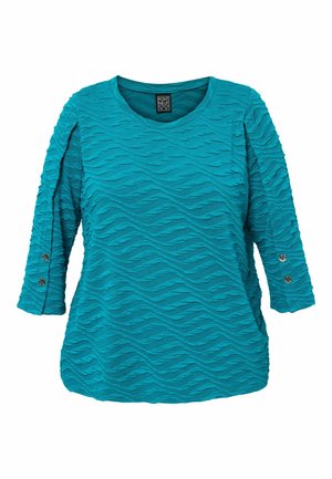 Teal-toppi, joka on valmistettu laadukkaasta kankaasta ja jossa on aaltoilevia kuvioita, kolmennesosa hihat ja napitusviyhetyksessä. Pyöreä kaula-aukko.