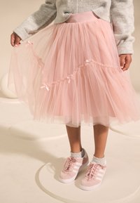 Ružová tylová sukňa s vrstvenými volánikmi, zdobená malými mašličkami. Kombinovaná s ružovými teniskami s bielymi pruhmi. Nad tým viditeľný sivý sveter.