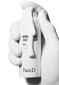 Bottiglia cilindrica bianca etichettata "CREMA HYDRABLAST 24H" tenuta in una mano guantata, con crema che fuoriesce dalla parte superiore, finitura liscia e opaca.