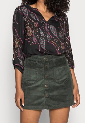 Kvinde iført sort paisley-mønstret bluse med pink, orange og grønne detaljer, kombineret med en mørkegrøn mini nederdel i kordfløjl med knapper.