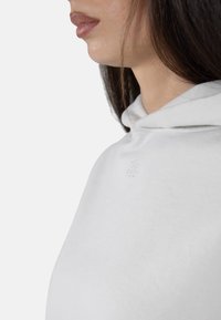 Lys grå hettegenser med en myk tekstur, som har en liten brodert logo på brystet og snor ved hetten. Nærbilde av krageområdet.
