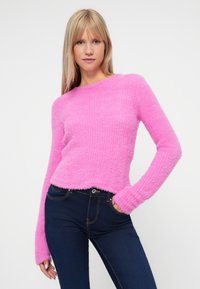 Puszysty różowy sweter z długimi rękawami, dekoltem w łódkę i skróconym dołem, w połączeniu z ciemnoniebieskimi dżinsami z przednimi kieszeniami. Widoczna miękka tekstura.