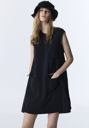 Frau trägt ein ärmelloses schwarzes Kleid mit Taschen und einen schwarzen, strukturierten Bucket-Hut, steht vor einem schlichten hellen Hintergrund.