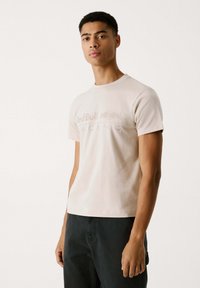 Lyserød bomulds-t-shirt med korte ærmer, rund halsudskæring og præget "Red Bull Racing" logo over brystet. Båret med mørke bukser.