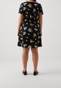 Robe noire à manches courtes, motif floral en rose et vert, taille ceinturée, ourlet au-dessus du genou, accompagnée de ballerines noires.