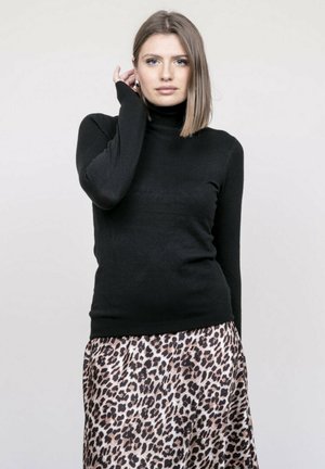 Jeune femme aux cheveux raides mi-longs portant un pull à col roulé noir et une jupe à imprimé léopard, posant avec la main près de l'oreille.