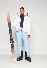 Sífelszerelés fehér pufi dzsekivel, tengerészkék felsővel, világoskék sí nadrággal és fekete síbakancsokkal, bal kézben egy sílécet tartva.