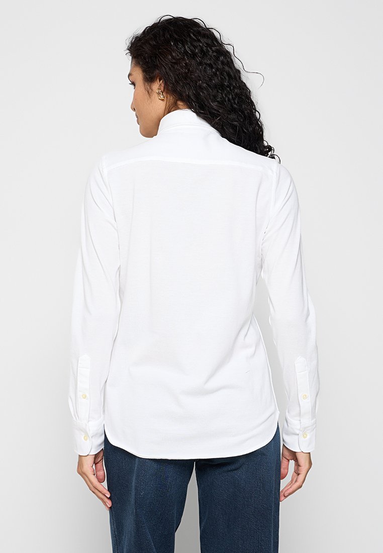 Camicia bianca a maniche lunghe con colletto e polsini con bottone, caratterizzata da una superficie liscia e un orlo dritto, vista da dietro.