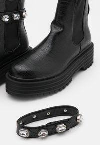 Czarne buty w krokodyli wzór z grubą podeszwą, uzupełnione dopasowaną czarną bransoletką z trzema przezroczystymi kamieniami szlachetnymi.