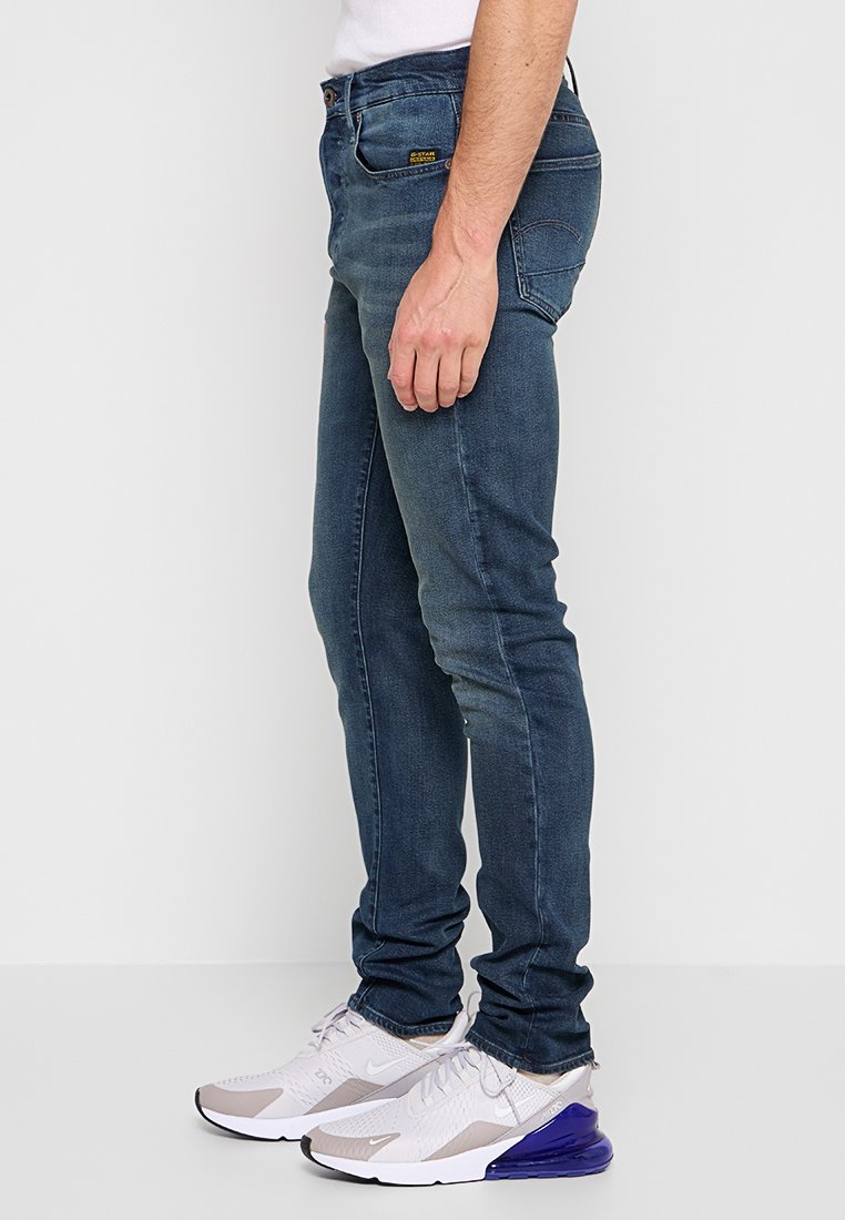 Jeans en denim slim-fit bleu foncé avec cinq poches, des détails de couture et une étiquette à la taille, associés à des chaussures de sport grises et blanches.