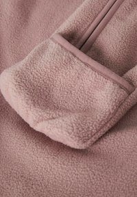 Zachte fleece stof in een gedempt stofroze met een opgerolde manchet die de textuur en afgewerkte naad toont.