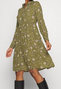 Langarmige, met kraag, jurk van olijfgroene stof, met een bloemenprint van roze en witte bloemen, taille-naad en een vloeiende rok.