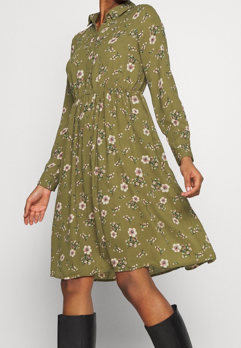 Langarmige, met kraag, jurk van olijfgroene stof, met een bloemenprint van roze en witte bloemen, taille-naad en een vloeiende rok.