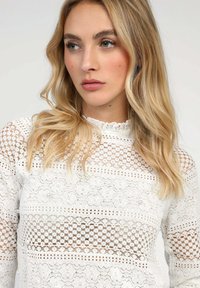 Top en dentelle blanche avec des motifs intriqués, présentant un col haut et des manches longues transparentes. Les détails incluent des motifs floraux texturés et des motifs en grille.