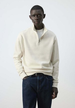 Hombre que lleva sudadera de cremallera corta color crema y vaqueros azul oscuro, de pie con una mano en el bolsillo contra un fondo liso.