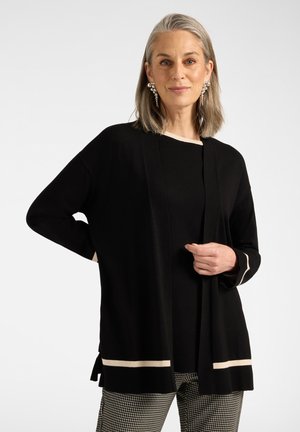 Frau mit grauen Haaren trägt einen schwarzen Cardigan mit weißem Besatz, ein schwarzes Top, Hahnentritt-Hose und große Perlenohrringe, steht mit der Hand in der Hüfte.