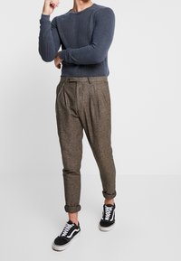 Maglione a coste navy, pantaloni in tweed marrone con pieghe e sneakers nere con accenti bianchi. Outfit casual con maniche arrotolate.