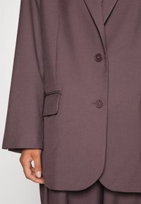 Hnědý přizpůsobený blazer s hladkou texturou, zapín�ání na jeden knoflík a kapsou na levém rukávu. Volný střih s zaobleným lemem.