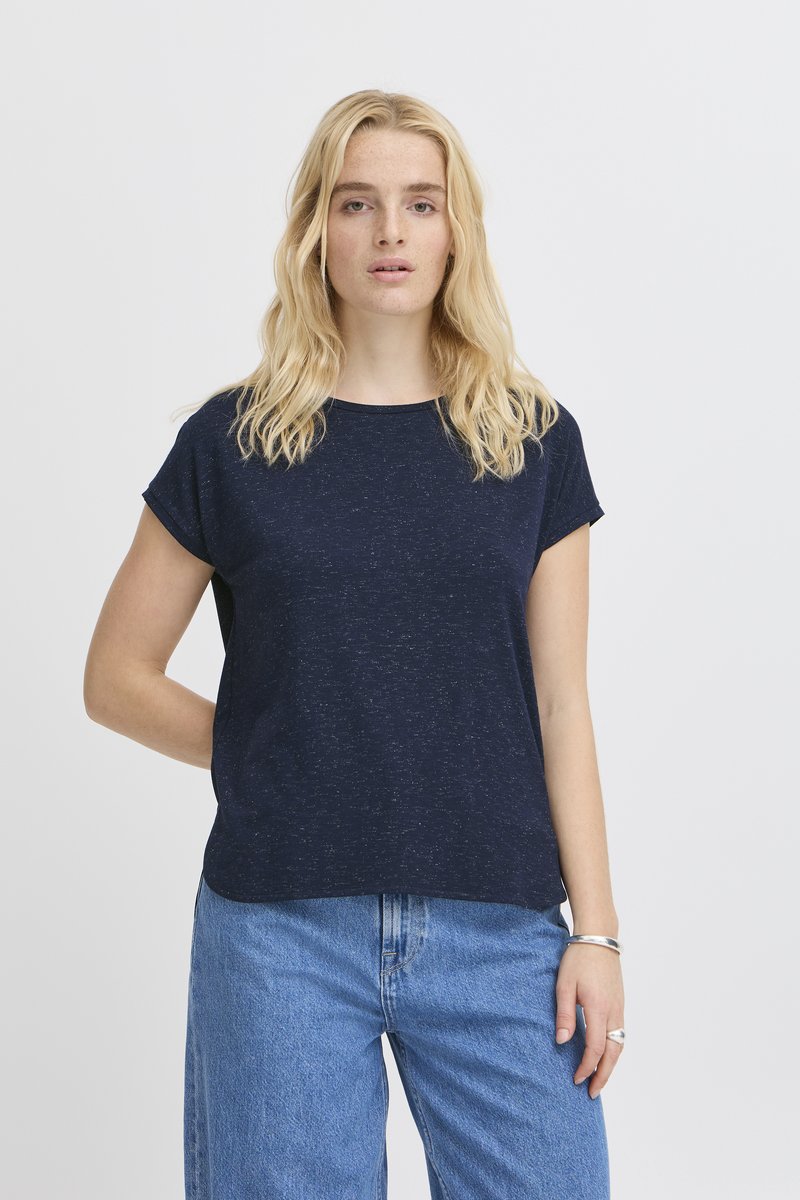 Femme blonde portant un chemisier bleu foncé à manches courtes et un jean bleu clair, debout devant un fond gris clair uni.