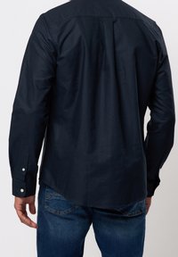 Granatowa koszula na guziki z długimi rękawami; płaski kołnierz, karczek z tyłu oraz zaokrąglony dół. Tkanina wygląda na gładką z lekko usztywnionym wyglądem.