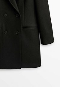 Manteau noir croisé avec quatre boutons visibles, deux poches fendues, manches longues et col cranté, présenté sur fond blanc.