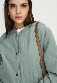 Veste matelassée vert clair avec un col rond, des boutons noirs et une sangle d'épaule marron, présentant un tissu texturé et une coupe décontractée.